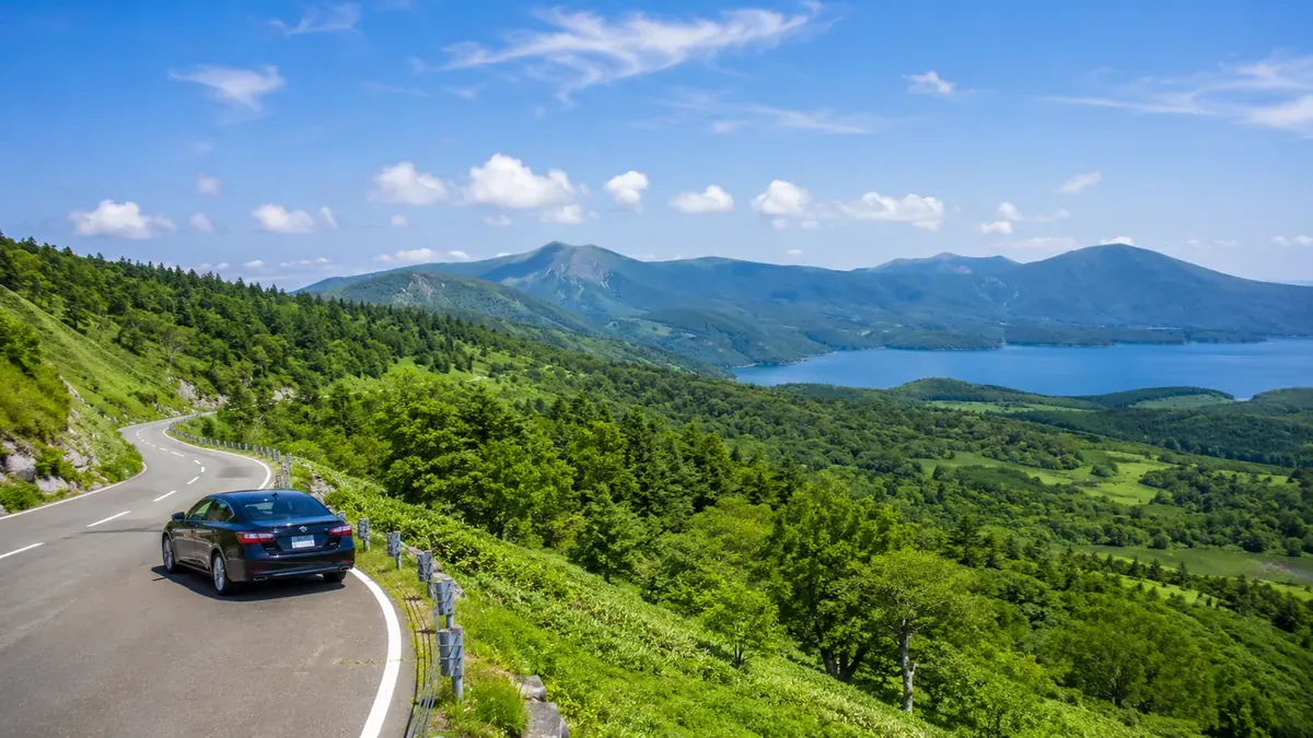 夏の青森の山々、湖、海岸線を背景に爽やかにドライブする一台の車